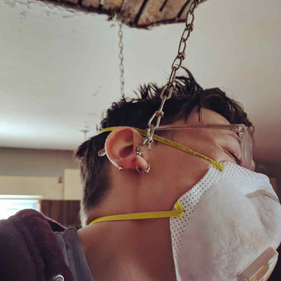 a close up of sadie, a butch woman wearing an n-95 mask, who has her earring caught on a drop ceiling chain
