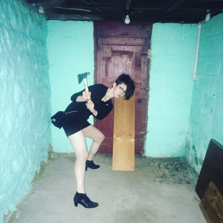 Nico is wearing a little black dress and heeled boots but is using a carpenter's axe to board a door shut