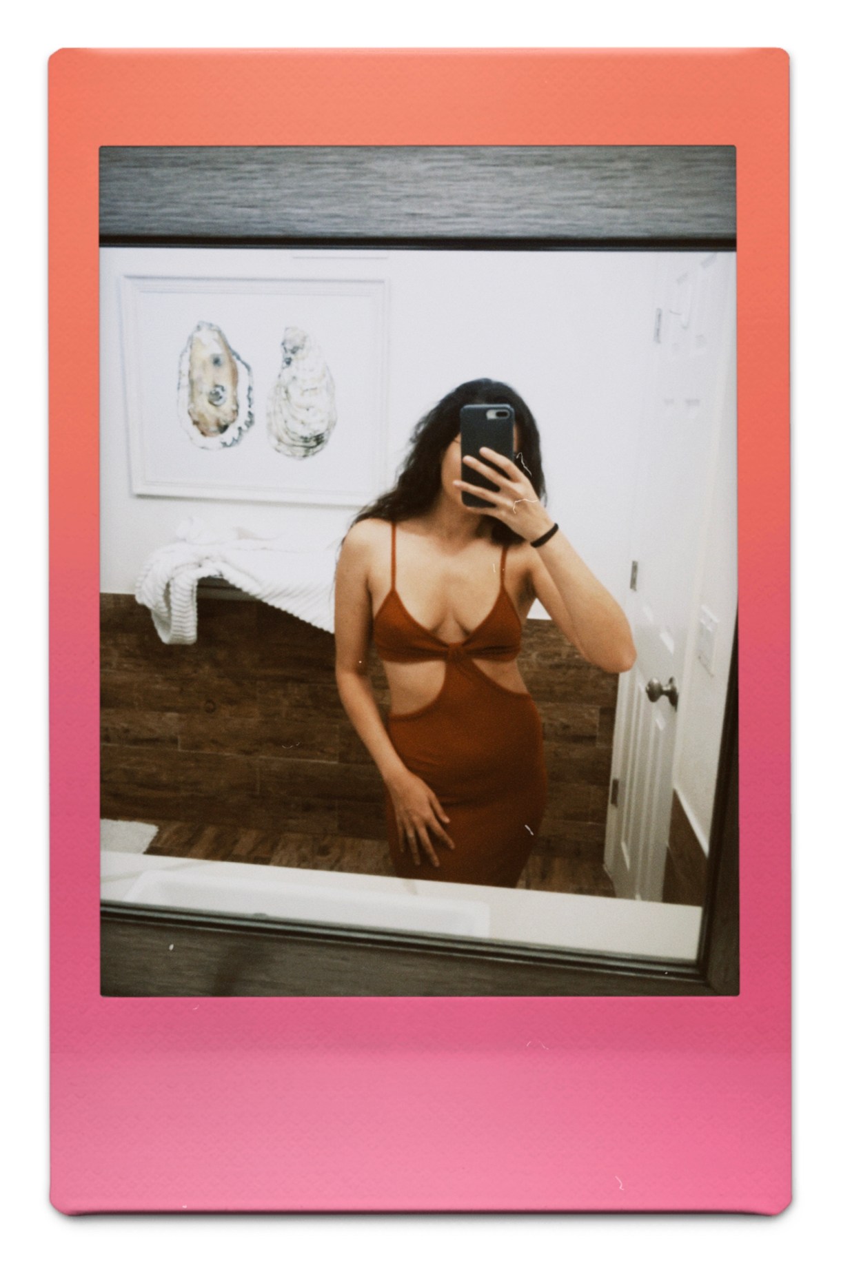 Kayla Kumari Upadhyaya takes a mirror selfie that covers her face but shows a brown spaghetti strap tight dress with cutouts on both sides of her rib cage, and her long brown hair is down behind her. Her bathroom's oyster painting is behind her.