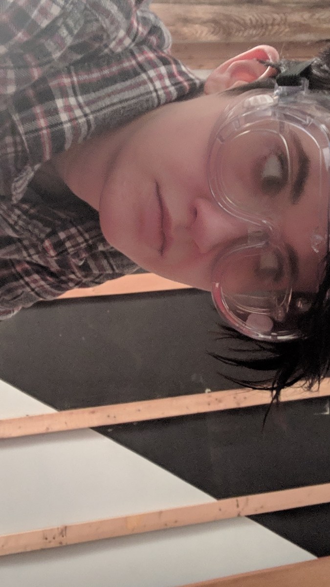 a photo of nico in a flannel and googles crouching in the foreground and giving side eye to a weird black triangle painted upside down and dominating a wall in the background