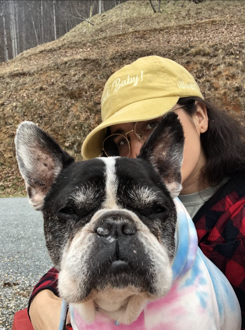 Kayla poses behind Lola a black and white French bull dog. Lola has cute puffy cheeks. Kayla is wearing a yellow baseball cap and red and black flannel.