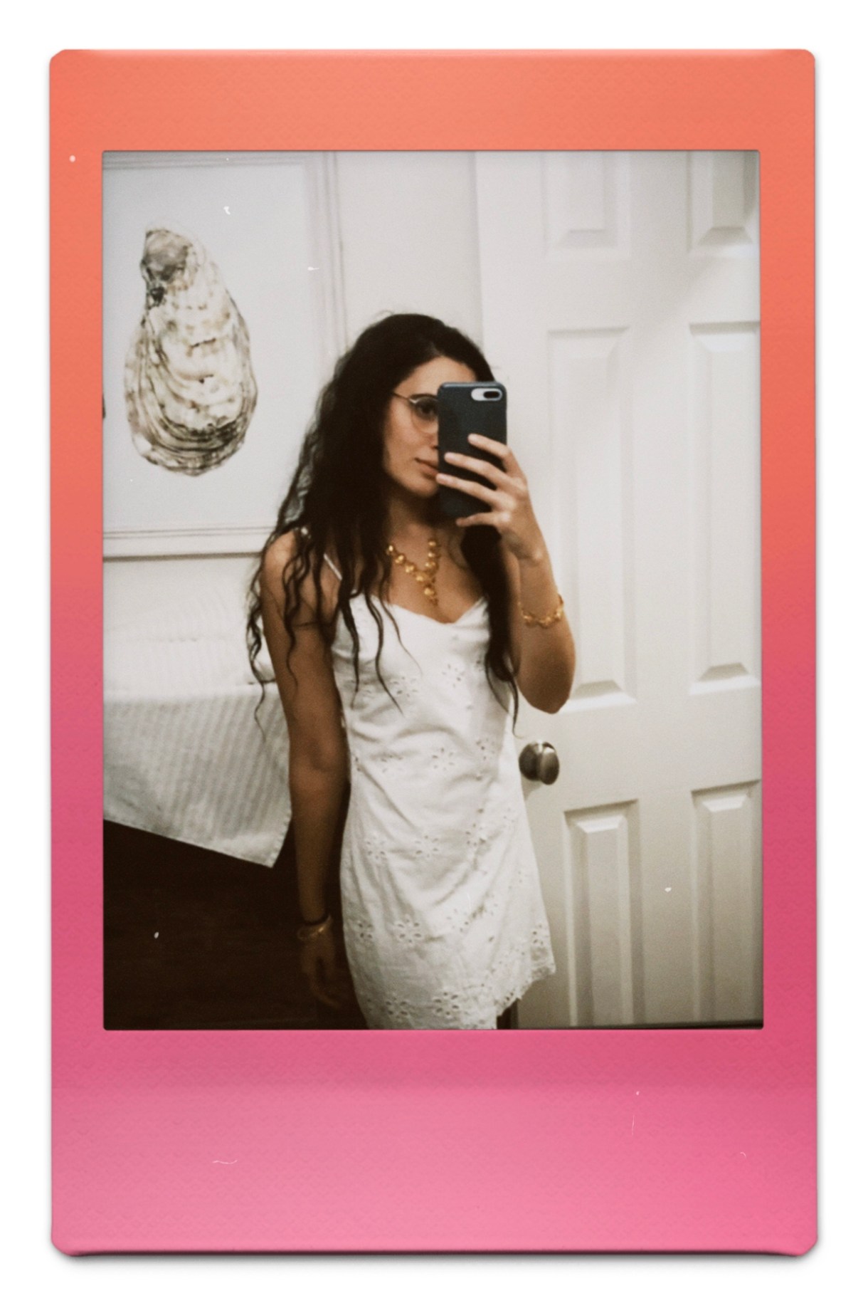 Kayla Kumari Upadhyaya wears an eyelet white dress and gold bangle and necklace while taking a mirror selfie in her bathroom. Her oyster painting is behind her.