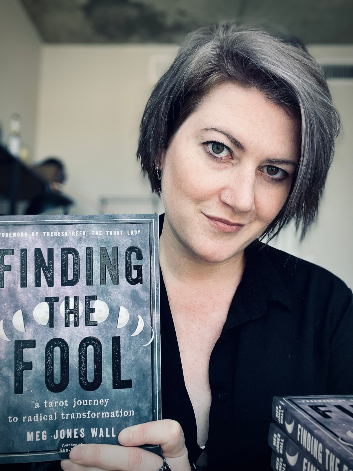 Meg, a white nonbinary human with gray hair holds their book "finding the fool" in front of them. they are wearing a black 