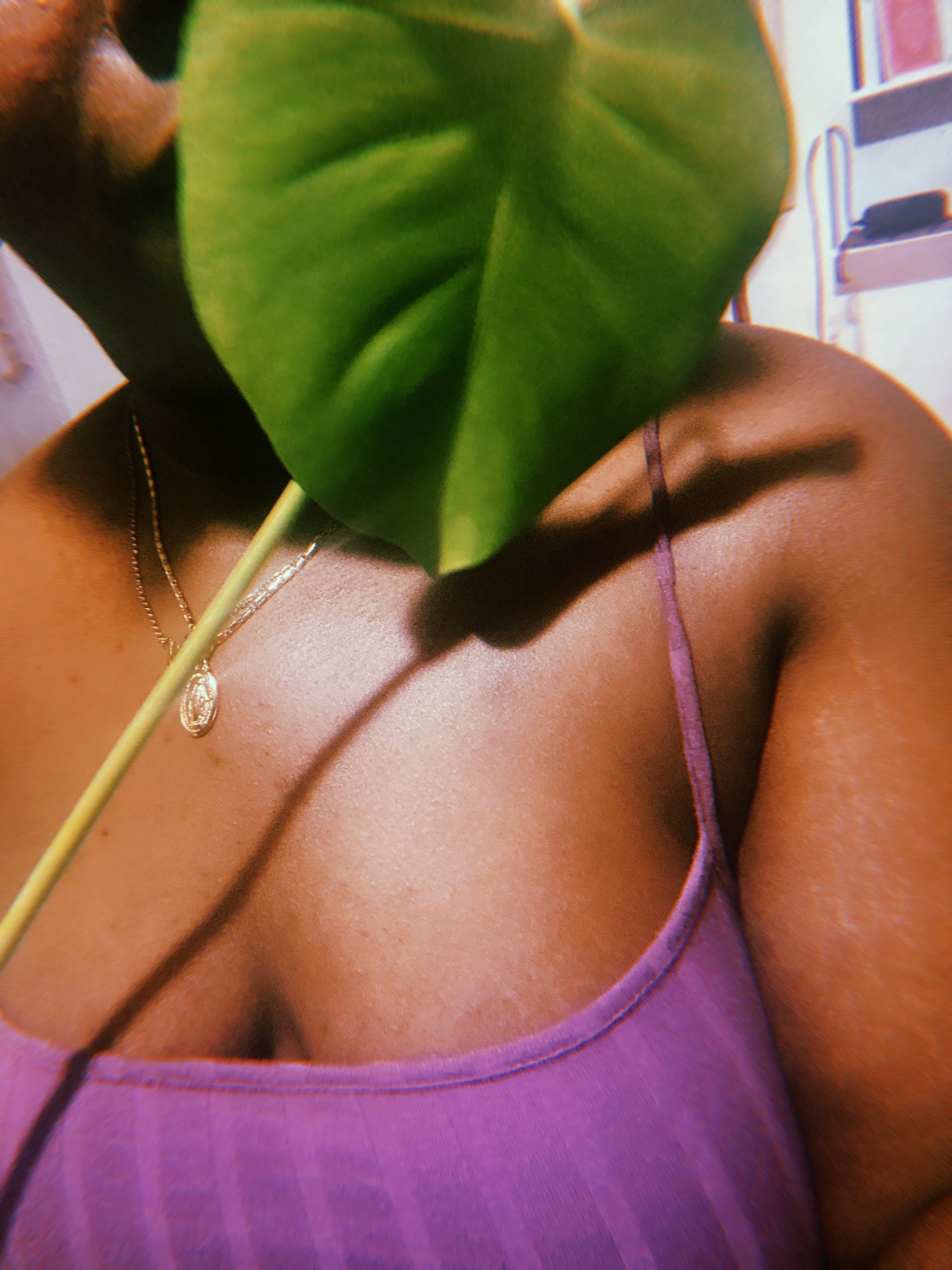 Shelli a Black woman sits behind a gorgeous plant leaf while wearing a purple camisole.