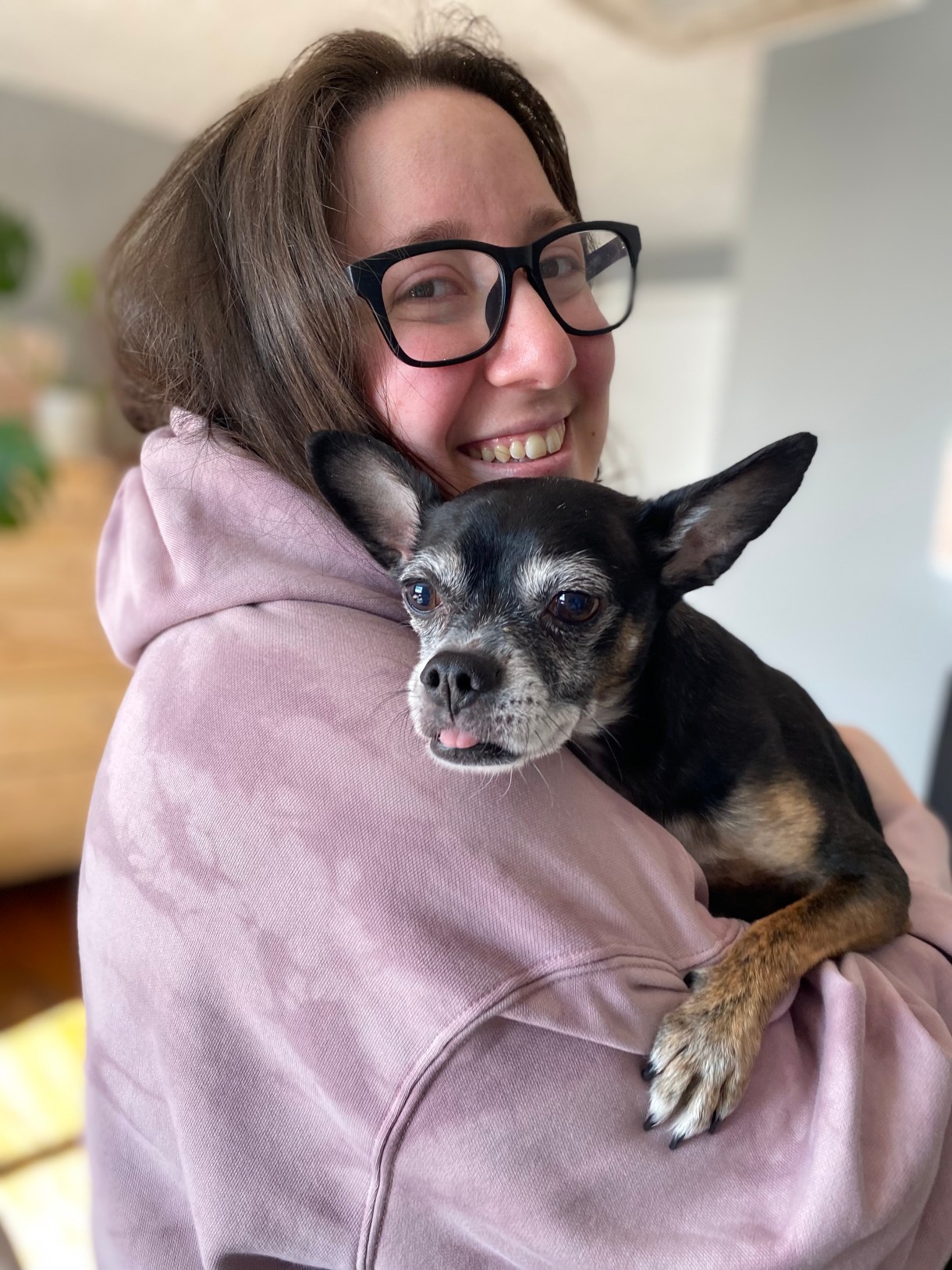 Vanessa smiles from behind a silly little black and white dog. Vanessa is a white woman with long brown hair and glasses. She is wearing a pink hoodie.