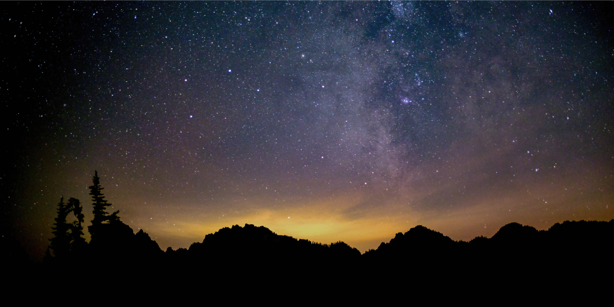 A blue and purple starry night sky is over shadow-covered trees and mountains. The sun is setting.