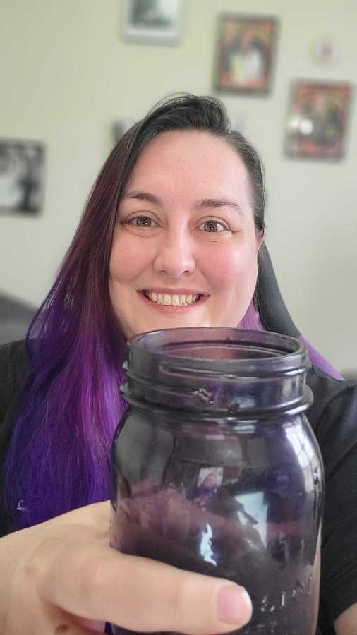 Valerie is a white woman with long hair dyed in a purple gradient. she is holding up a mason jar that is also purple to the camera and smiling