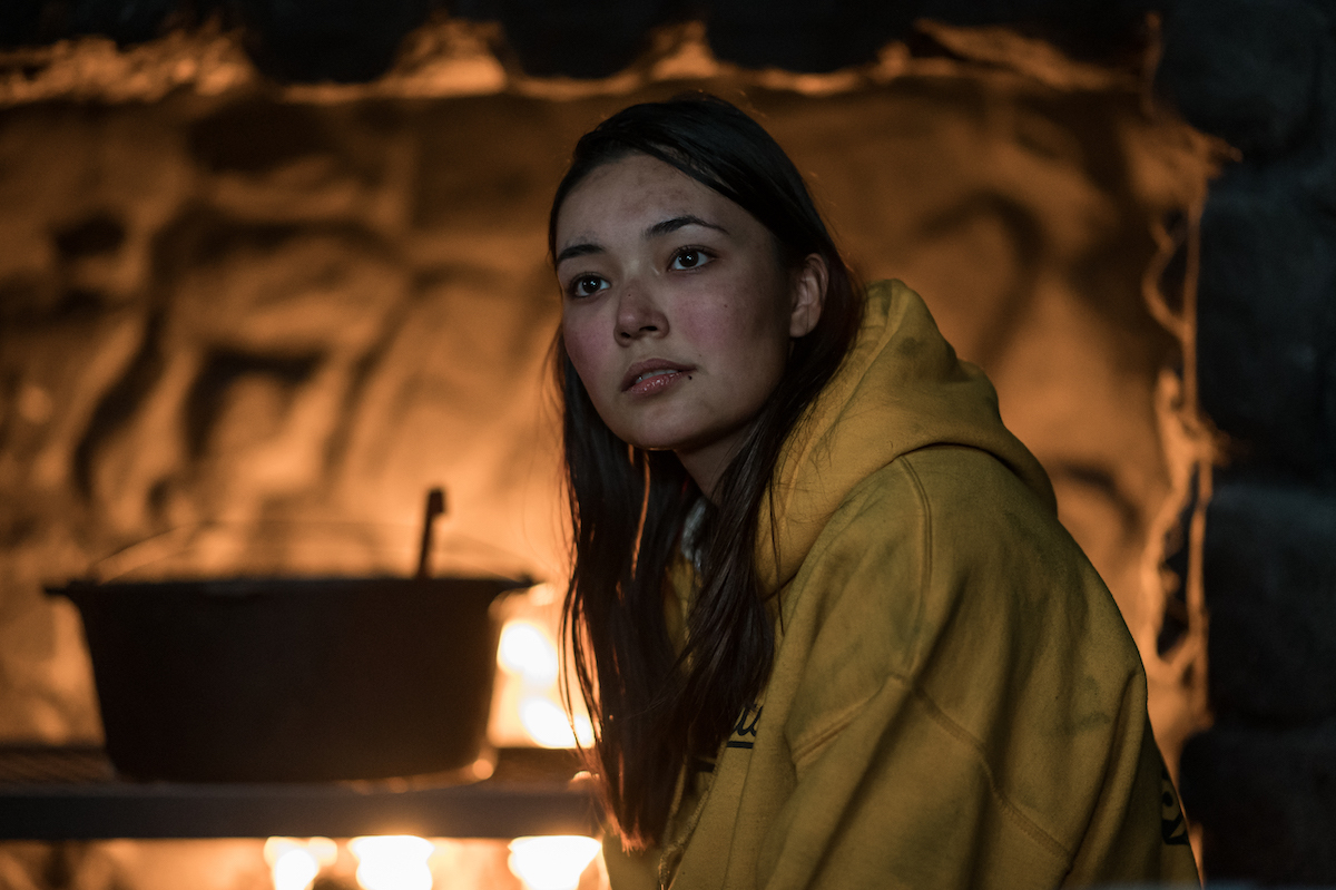 Yellowjackets Alexa Barajas as Teen Mari in YELLOWJACKETS Season 2 wears a yellow hoodie near a fireplace. Photo Credit: Kailey Schwerman/SHOWTIME.