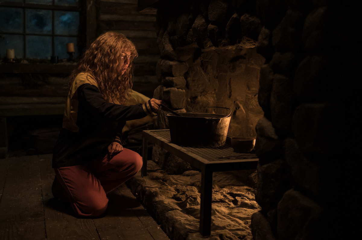 Yellowjackets Samantha Hanratty as Teen Misty in YELLOWJACKETS Season 2 kneeling in front of a fire cooking in the cabin. Photo Credit: Kailey Schwerman/SHOWTIME.