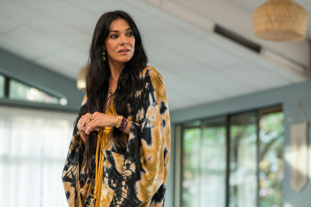 Simone Kessell as Lottie in YELLOWJACKETS Season 2 in a floral robe stands in sunlight. Photo Credit: Kailey Schwerman/SHOWTIME.