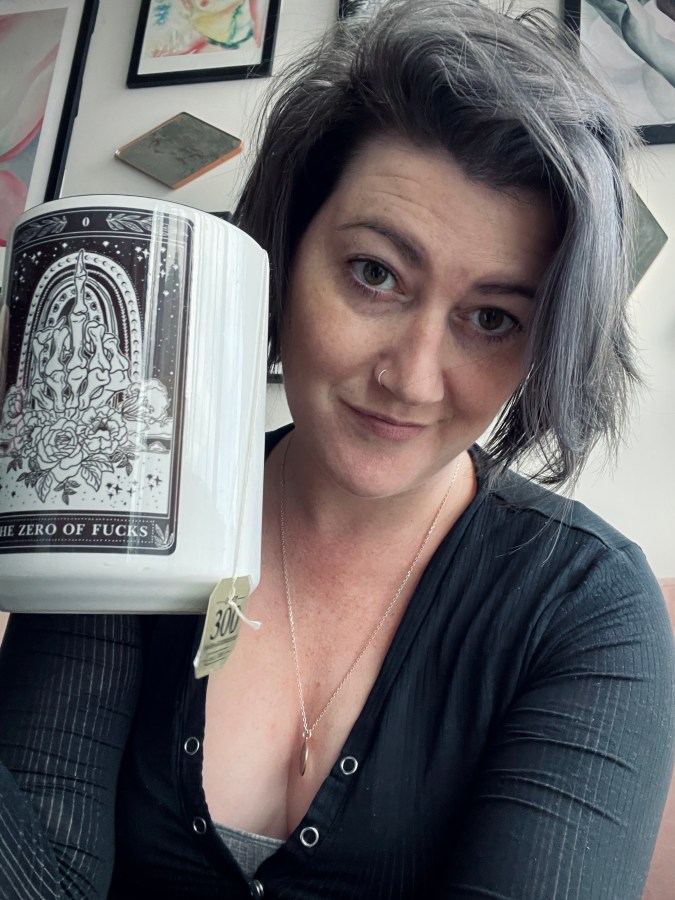 Meg is a white person with bobbed black and gray hair who is wearing black and holding up a tarot mug with tea to the camera.