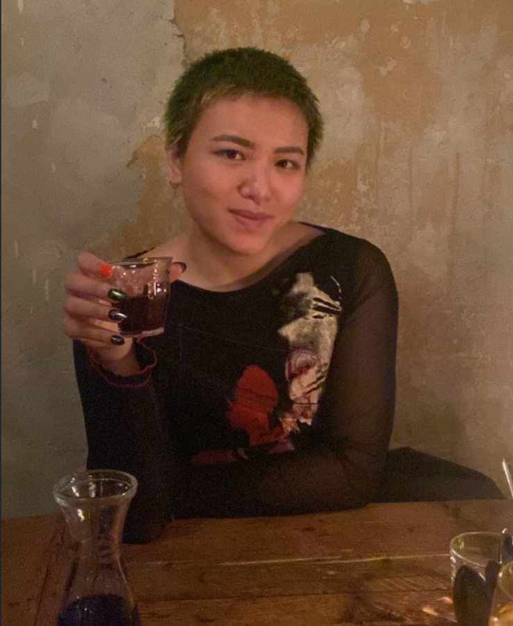 Viv, an Asian nonbinary person with short green hair is seated in a bar in Berlin holding a glass of red wine and smiling mischieviously. they are wearing an artsy black and abstractly patterned shirt.