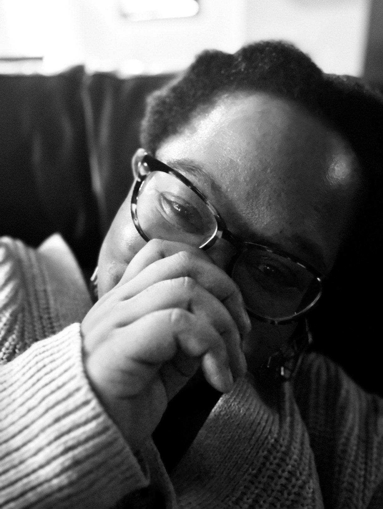 a black and white photo of carmen, a Black woman, smiling with her hand over her mouth, wearing a sweater and glasses. her hair is short