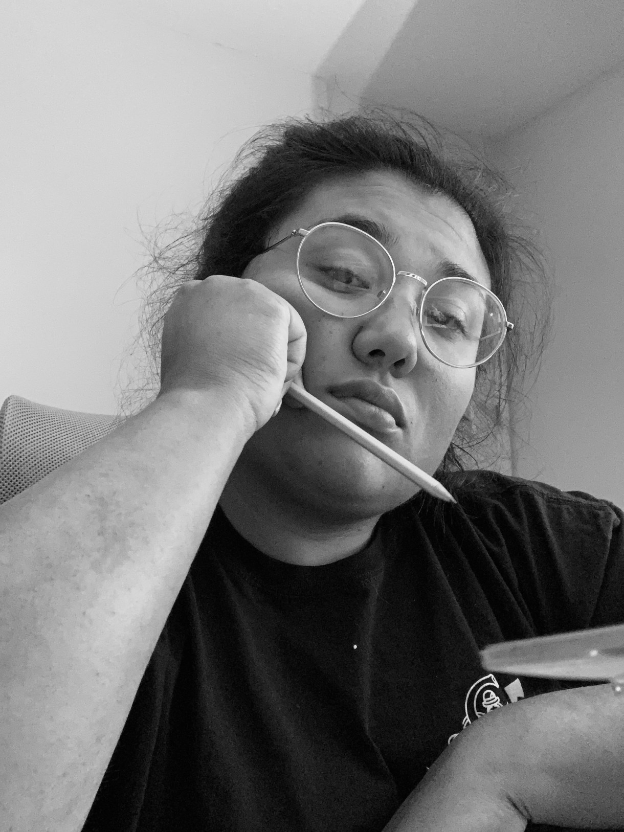 a black and white photo of Em, a mixed race person, holding a pencil and looking moody. she is wearing glasses.