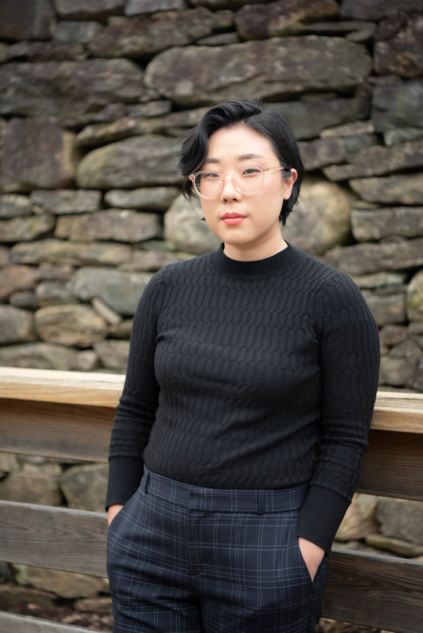 portrait of Franny Choi, an Asian American woman with short black hair wearing glasses, looking at the camera with a small smirk on her face. She is wearing a black long sleeve shirt and black pants and her hands are in her pockets.