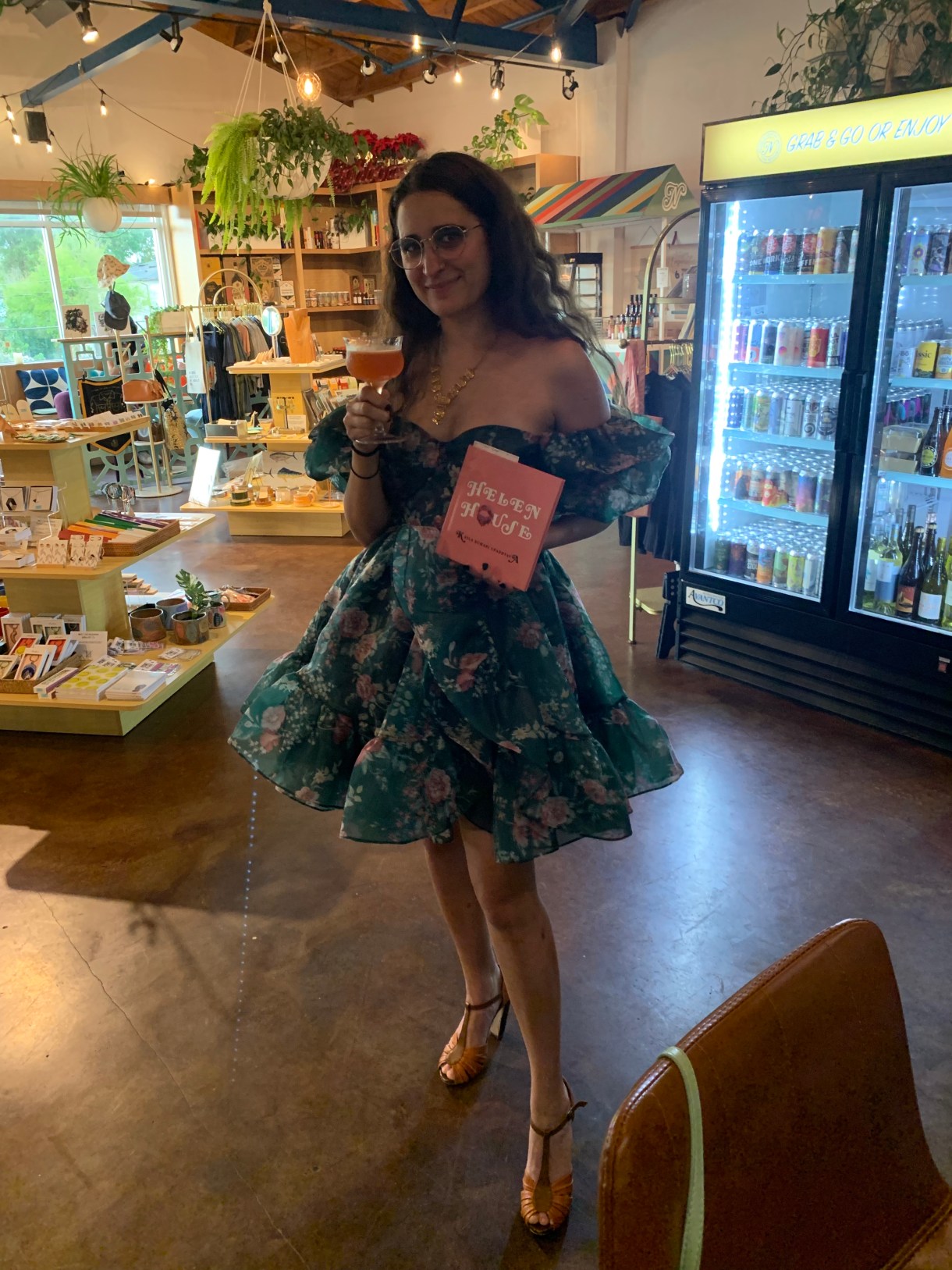 Kayla Kumari Upadhyaya in a green and pink floral dress holding her pink book HELEN HOUSE in one hand and a pink cocktail in the other.