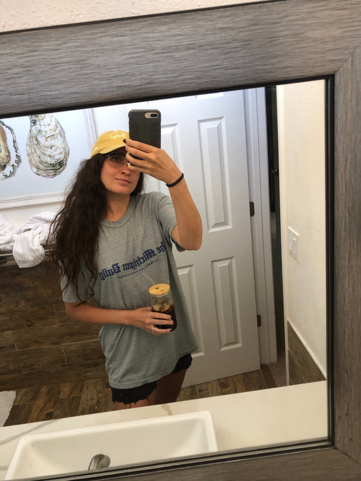 Kayla Kumari Upadhyaya in a gray tee that says THE MICHIGAN DAILY, a hat that says FERNET BABY, and black jean ripped shorts. She is holding a glass tumbler full of iced coffee and taking a mirror selfie.