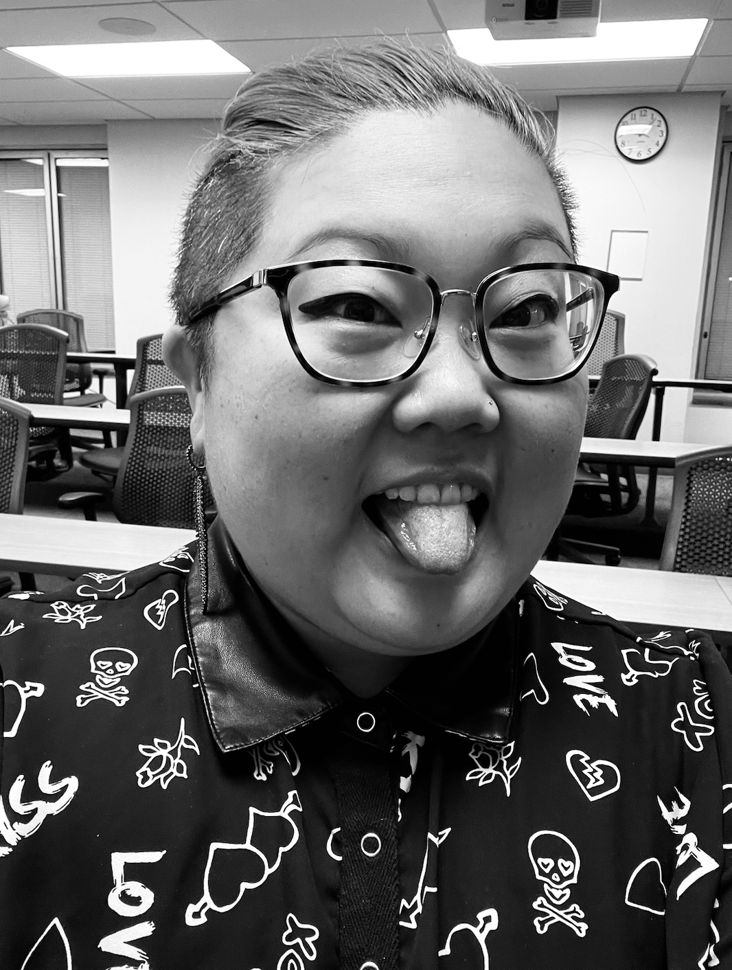 a black and white photo of KaeLyn an Asian woman who here is wearing a fun button up with graphic designs as well as glasses. She is sticking out her tongue. Her hair is pulled back.