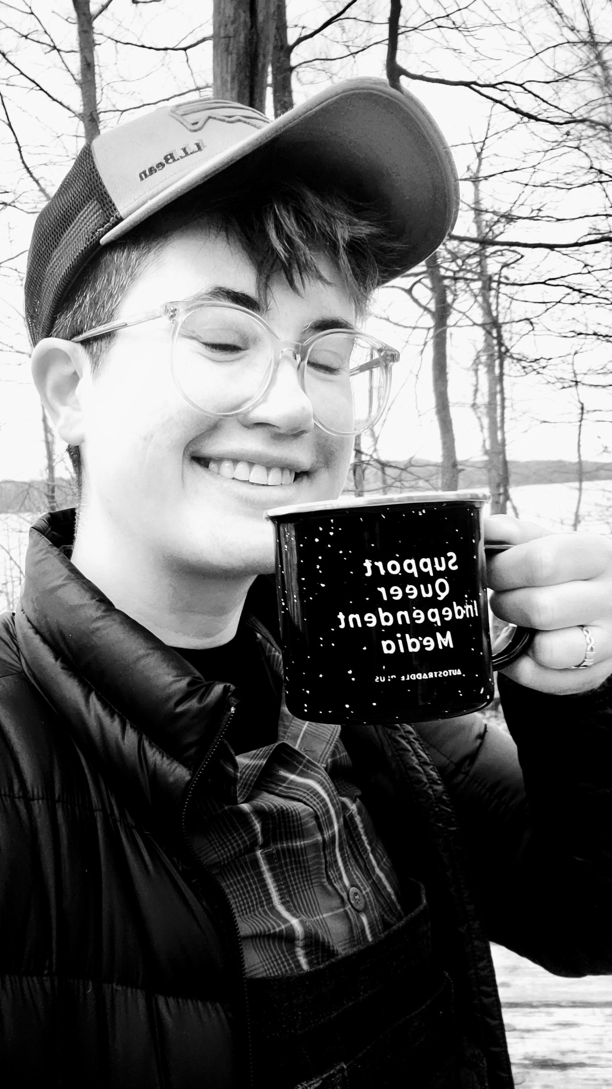 a black and white photo of nico, a white genderqueer human, drinking out of an A+ enamel cup in the woods, wearing a puffy coat, plaid shirt, trucker hat, and clear glasses