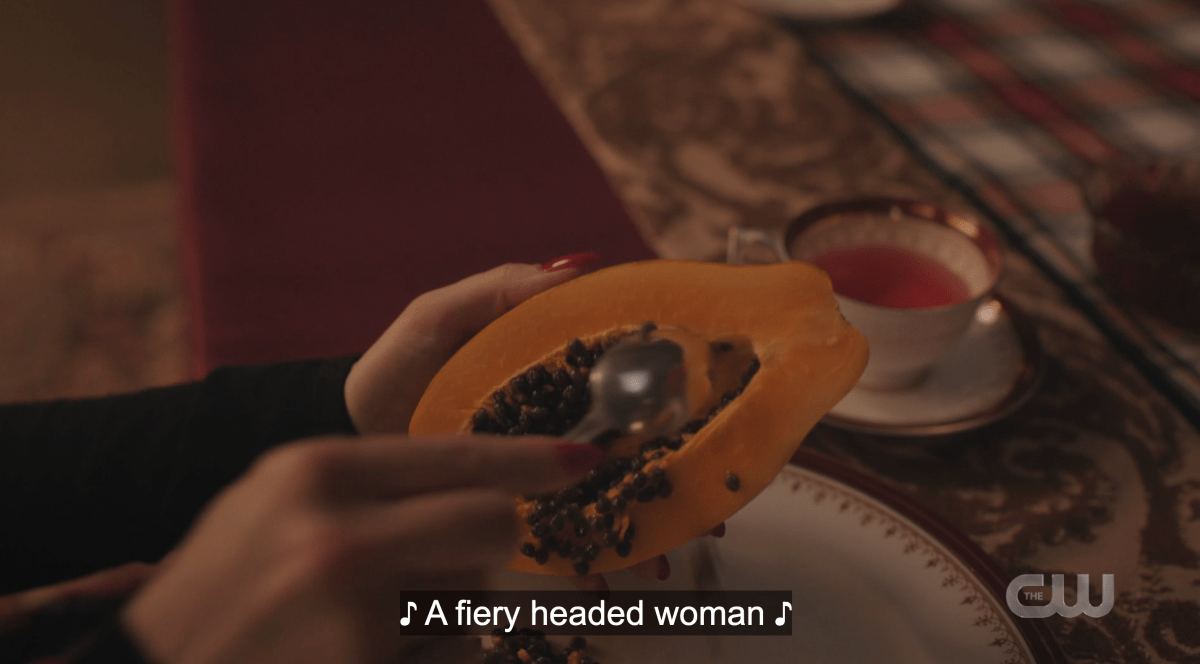 Cheryl scoops out papaya while "a fiery headed woman" is sung over top it