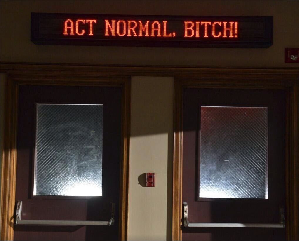 a marquee sign above two school doors that says in bright red letters "Act Normal, Bitch!"