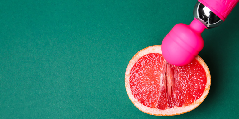 Against a teal background, half of a grapefruit is in the bottom right corner of the image. A hot pink wand vibrator extends from the upper right corner and rests against the grapefruit.