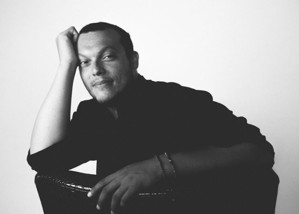 portrait of Charif Shanahan, a light skinned black man with short cropped black hair, leaning on his right fist in a comfortable gesture as he softly looks at the viewer. He is wearing a black top and has two bracelets on their left wrist.