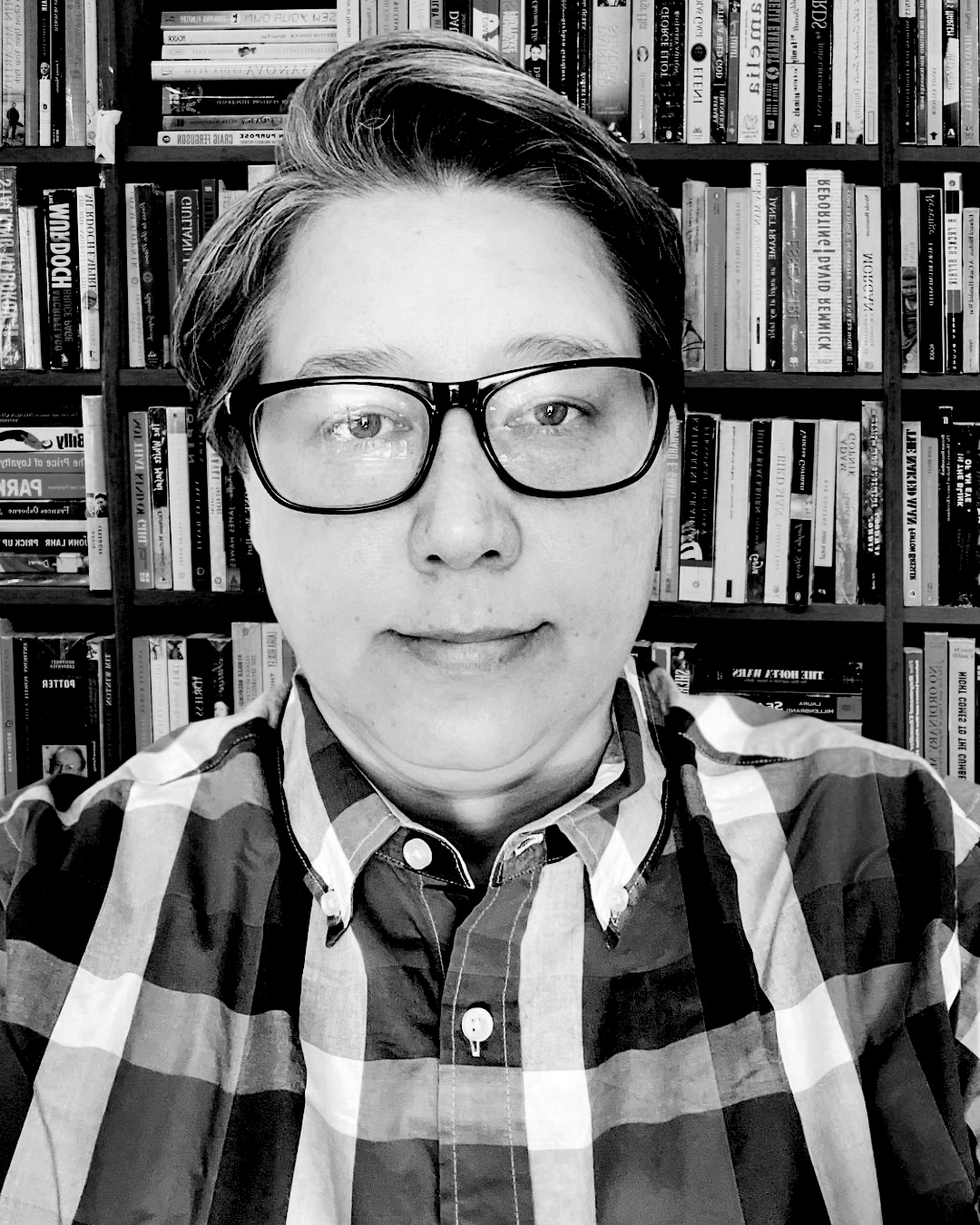 a black and white photo of Heather a soft butch woman with glasses wearing a plaid button up shirt and looking serious