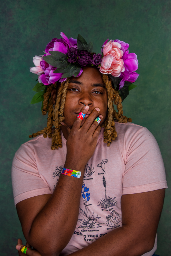 portrait of kb brookins, a black trans person with a flower crown, holding their face where multi colored band-aids can be seen on their hands and wrist. They are wearing a pink top and looking at the viewer. They have golden shoulder length twists.