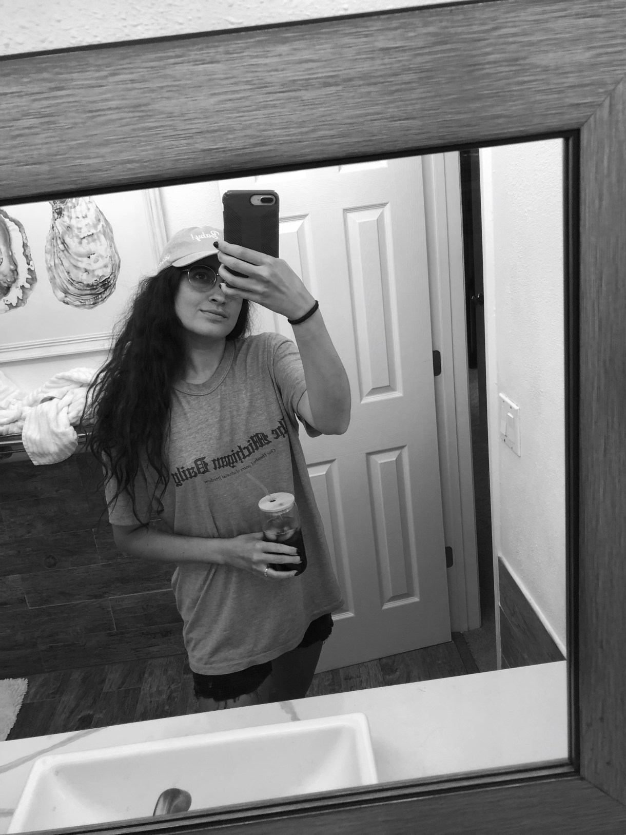 a black and white photo of Kayla, a South Asian woman, taking a selfie in a mirror. she is wearing a long tee and baseball cap and holding a beverage. her hair is long and wavy.