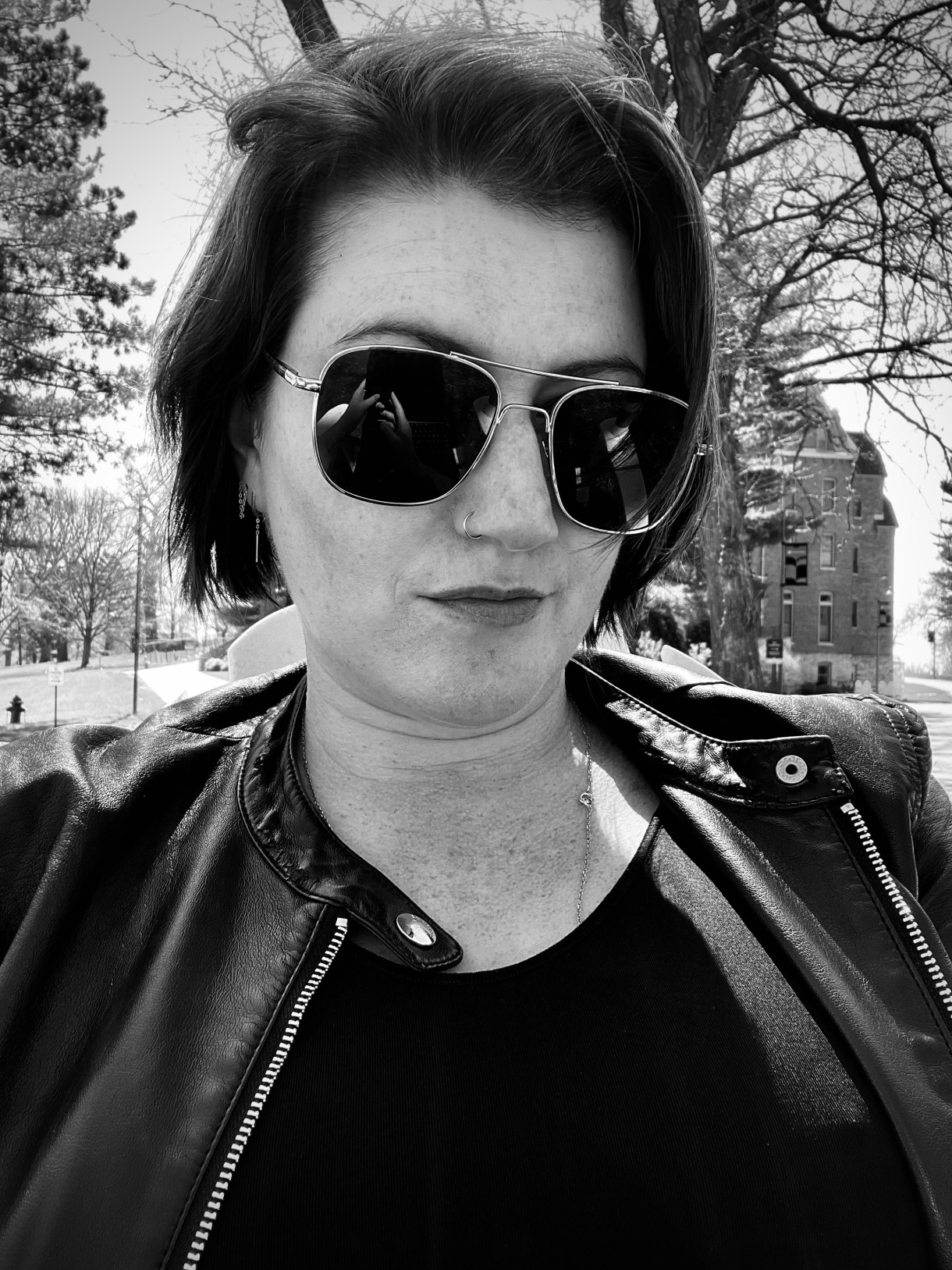 a black and white photo of meg a white person wearing sunglasses and a leather jacket. she has short dark hair.