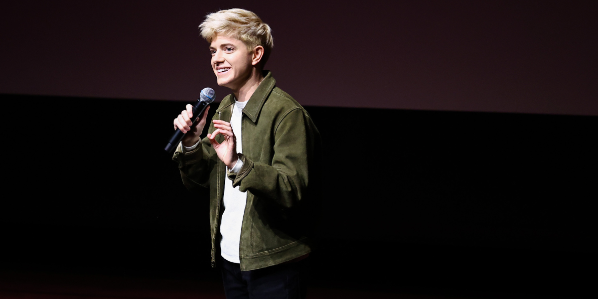 Mae Martin on stage with a mic wearing a white tee and an olive green open shacket