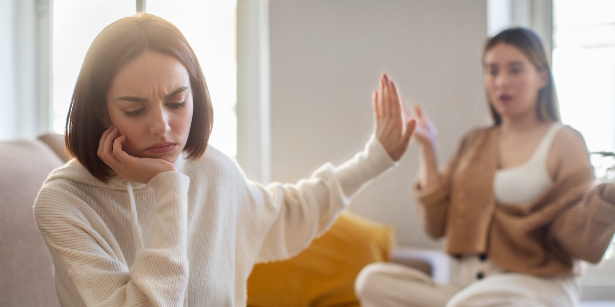 Two women are having an argument. One looks like she is trying to talk, while the other turns away and puts her hand up, signaling her to stop and that she doesn't want to listen.