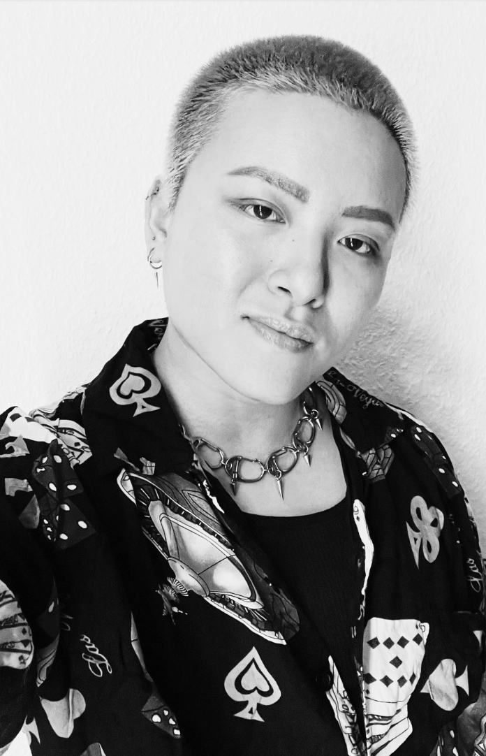 a black and white photo of viv, an Asian nonbinary human, who has short shaved hair and is wearing a coat patterned with cards and playing card symbols