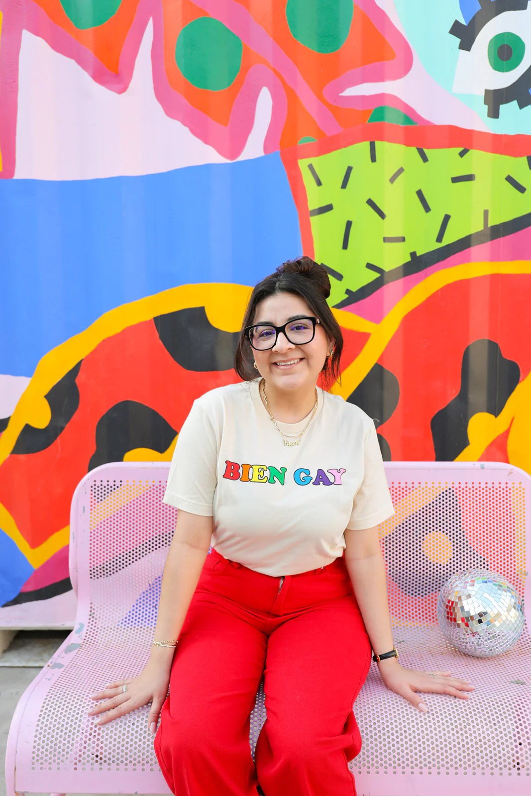 Girl smiling in glasses with red pants and a t-shirt that says BIEN GAY in rainbow lettering