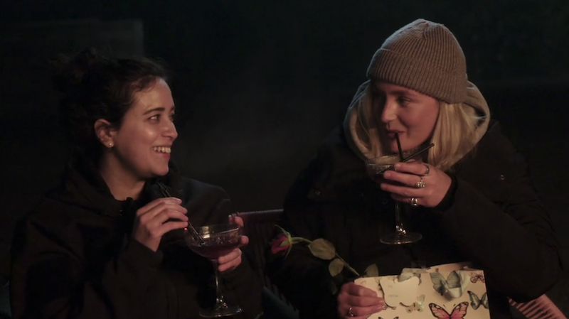 Statler and Dempsey sitting next to each other by a campfire; Statler is holding a drink and smiling at Dempsey who is sipping on her drink