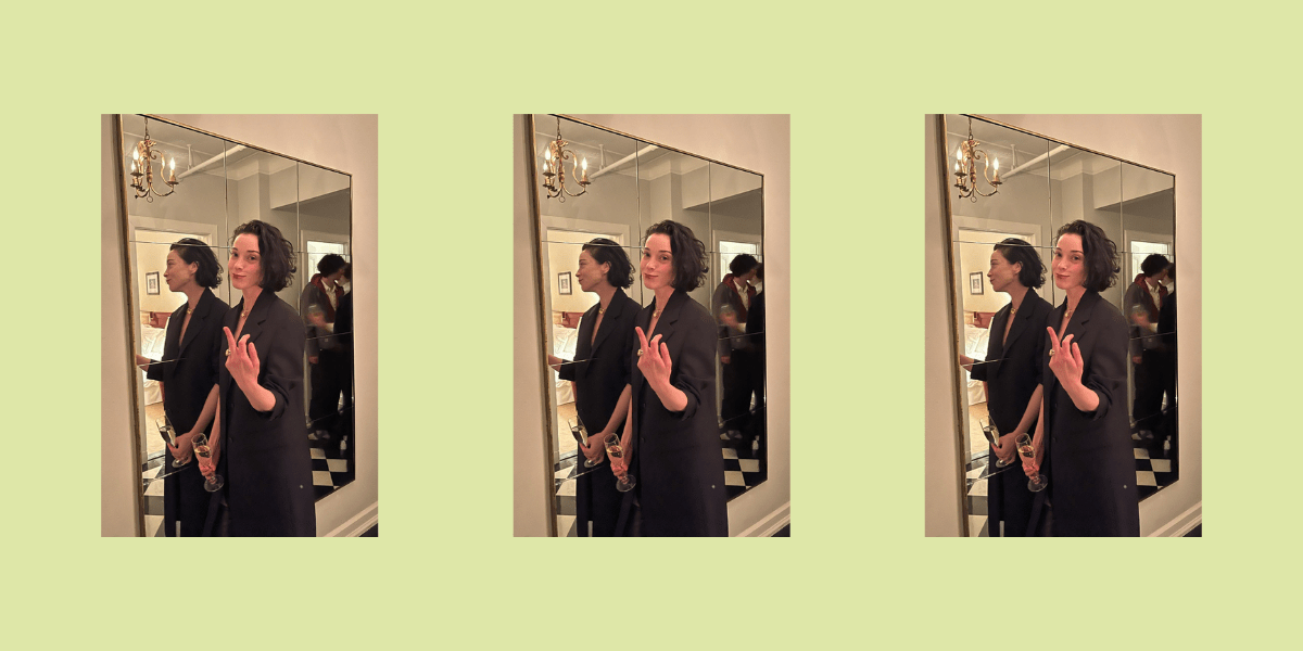 St. Vincent wearing a long black oat, throwing up a middle finger next to a mirror in a hallway