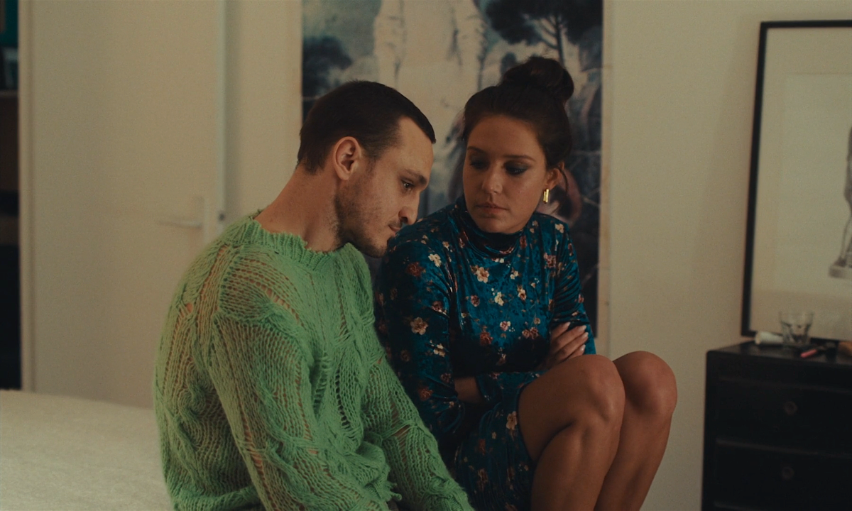 Franz Rogowski in a light green knit sweater sits on a bed next to Adèle Exarchopolous in a blue velvet dress.