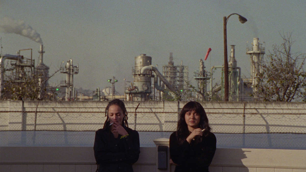 Best queer movies of 2023: Sasha Lane and Ariela Barer smoke cigarettes with a toxic waste plant blowing fumes behind them.