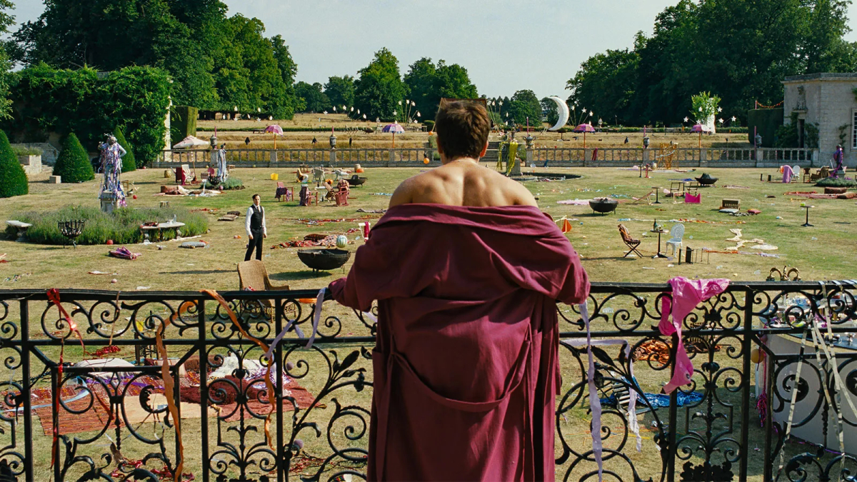 From behind a shot of Barry Keoghan with a red robe half on looks out over the aftermath of a party on the front lawn of a manor. 