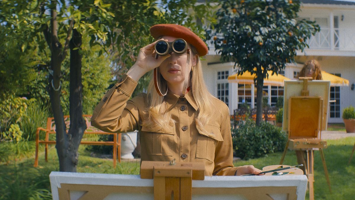 Anna Konkle in a beige leather jumpsuit and orange beret holds a pair of binoculars up to her face.