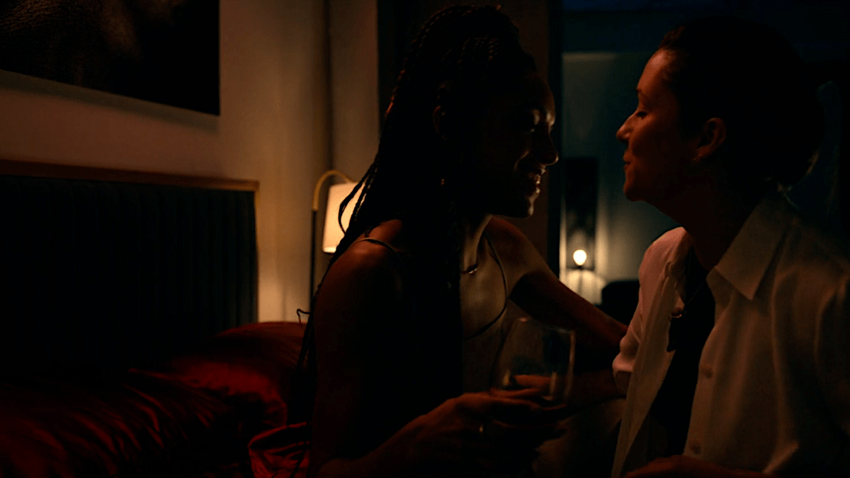 Two women shrouded in darkness smile at each other while sitting on a bed.