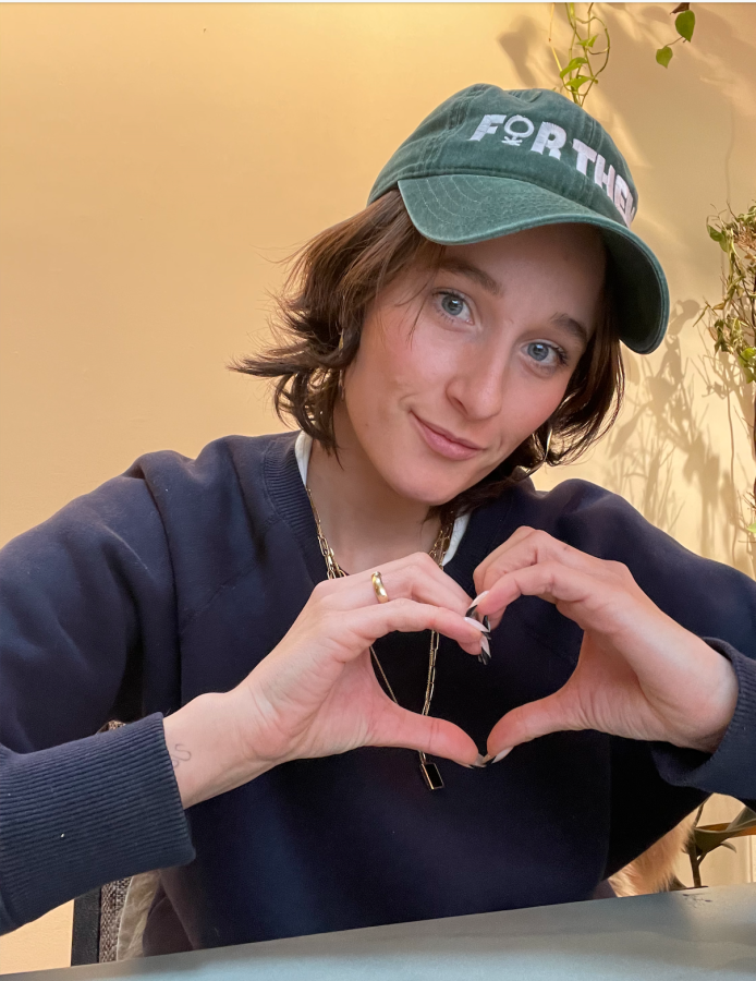 Em, a white woman with short wavy brown hair wearing a for them baseball cap holds her hands up in a heart shape