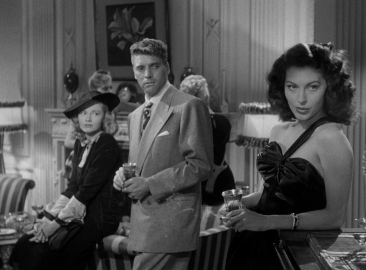 Ava Gardner leans on a bar as a man and woman watch her.