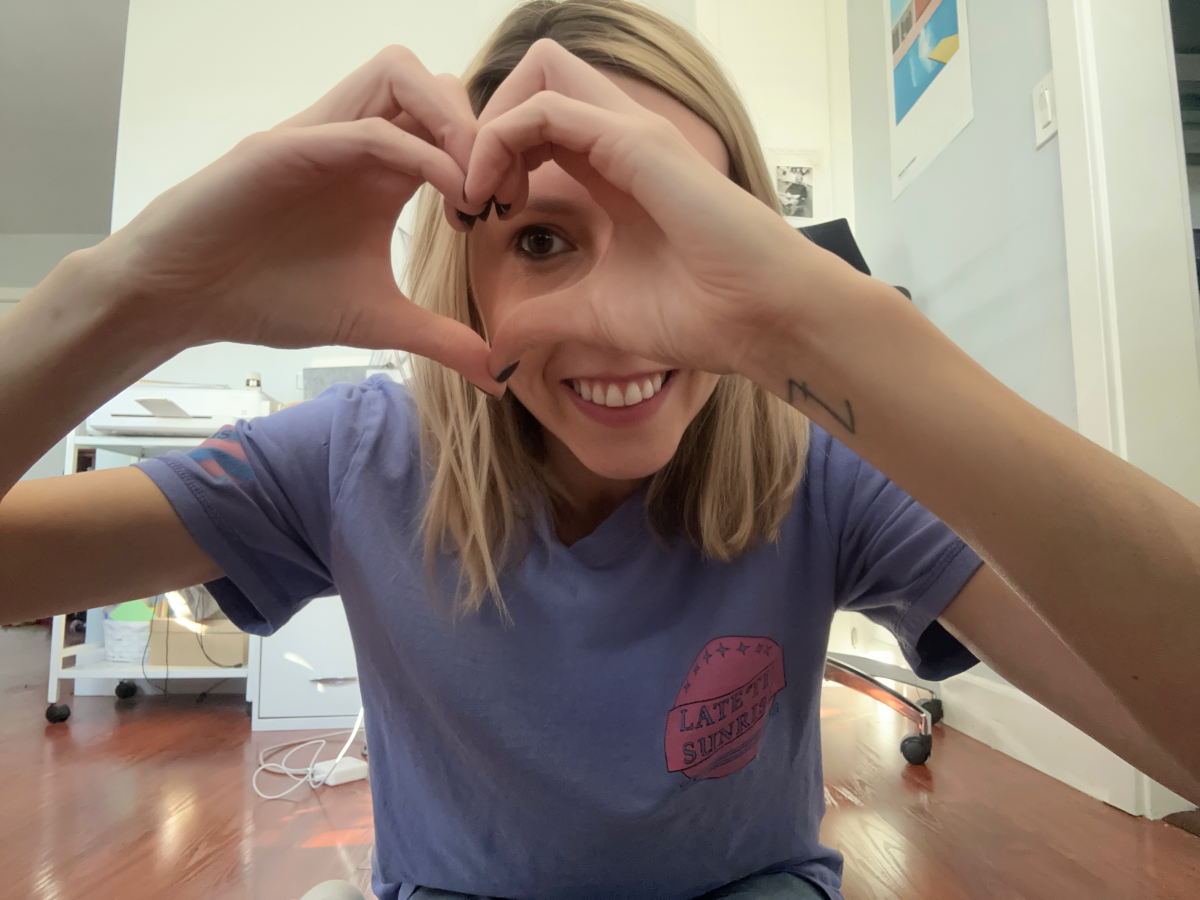 riese a white agender human with a blonde bob holds their hands up in a heart shape and peers through the center of the heart