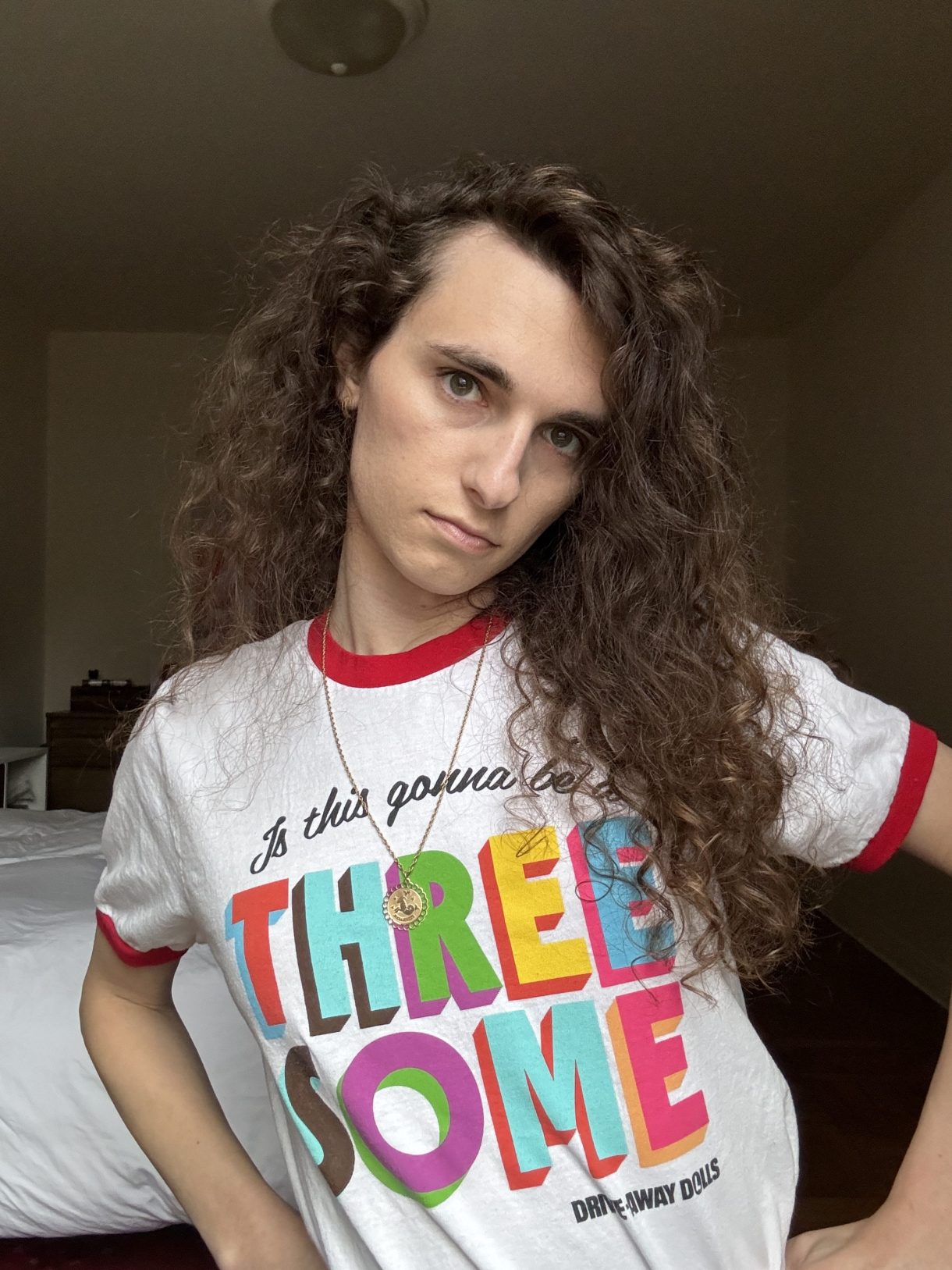 Drew is a white woman with long curly brown here. Here, she is posing with her hands on her hips while wearing a shirt that says "threesome" in huge colorful letters