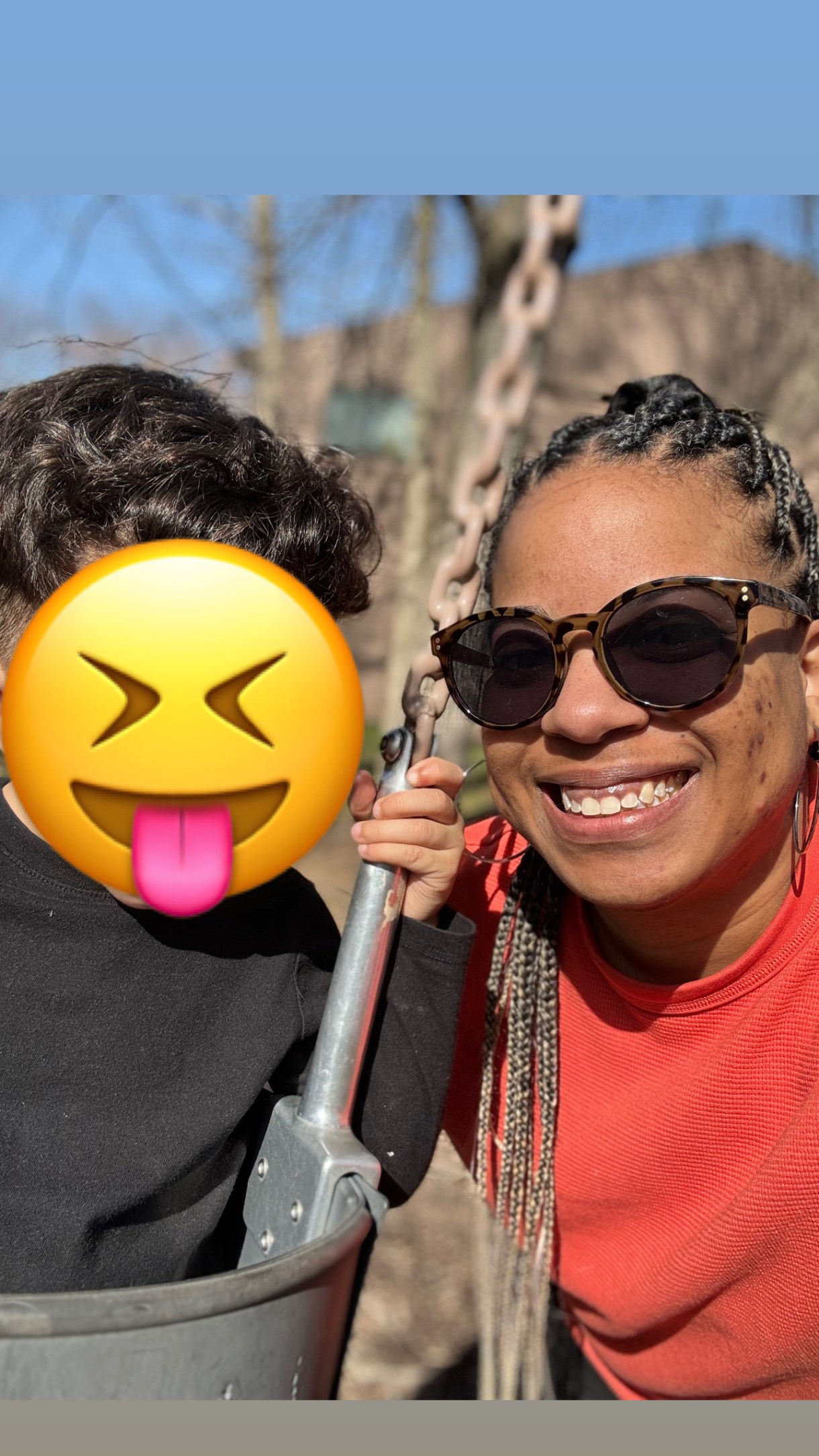 a photo of Nic a Black woman with long braids and sunglasses who is smiling outdoors in what looks like a park. She is wearing an orange sweatshirt.