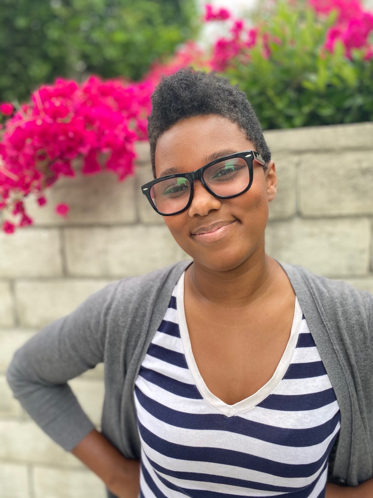 a photo of Sa'iyda who is a Black woman who is smiling and standing in front of pink flowers and greenery. She is wearing a blue and white striped shirt and cardigan. She has short curly hair and glasses.