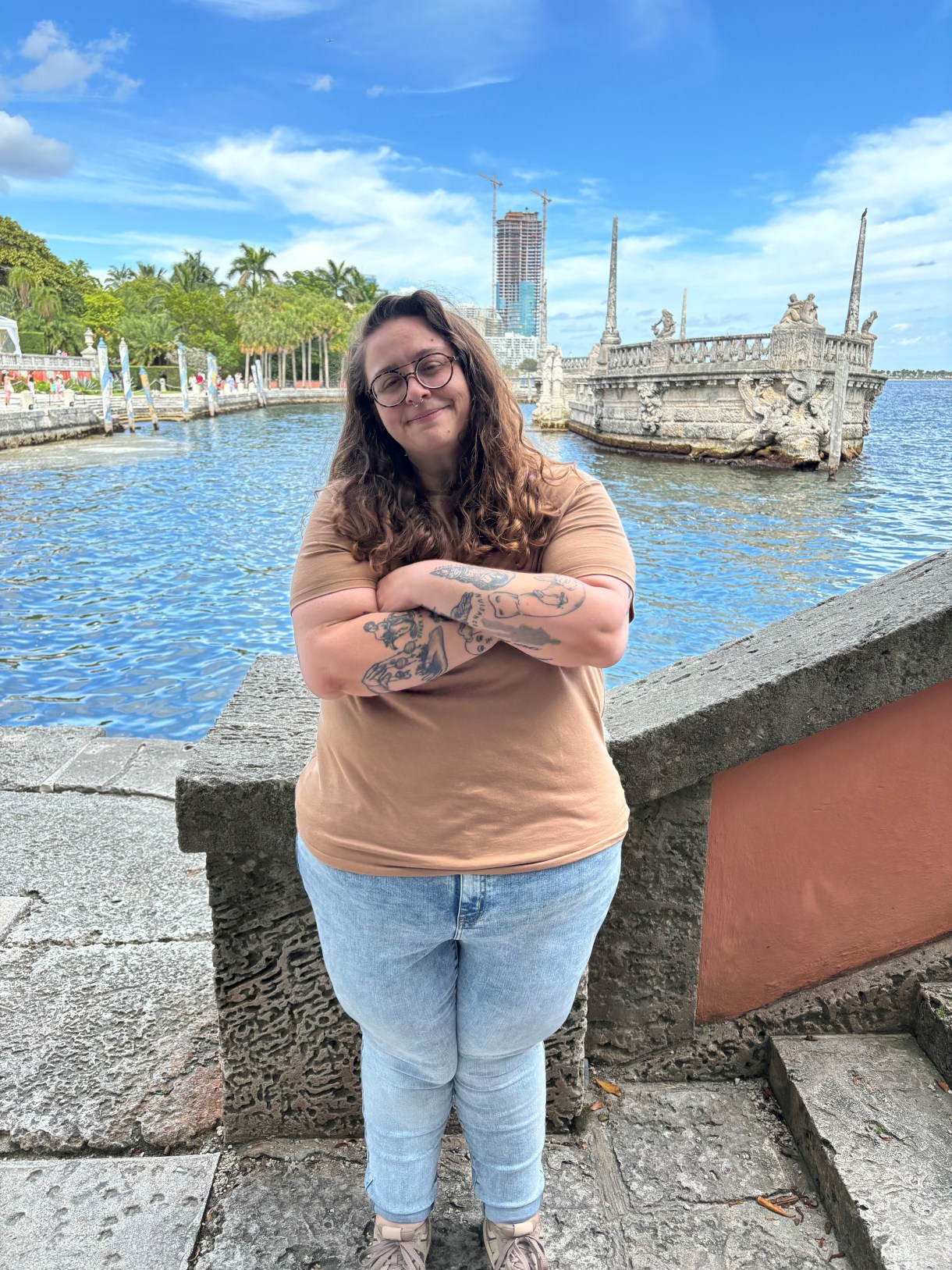 a photo of Stef a white nonbinary person standing with arms crossed in front of a body of water with a boat behind them. stef's arms are crossed and they are smiling, they have long brown hair and glasses.