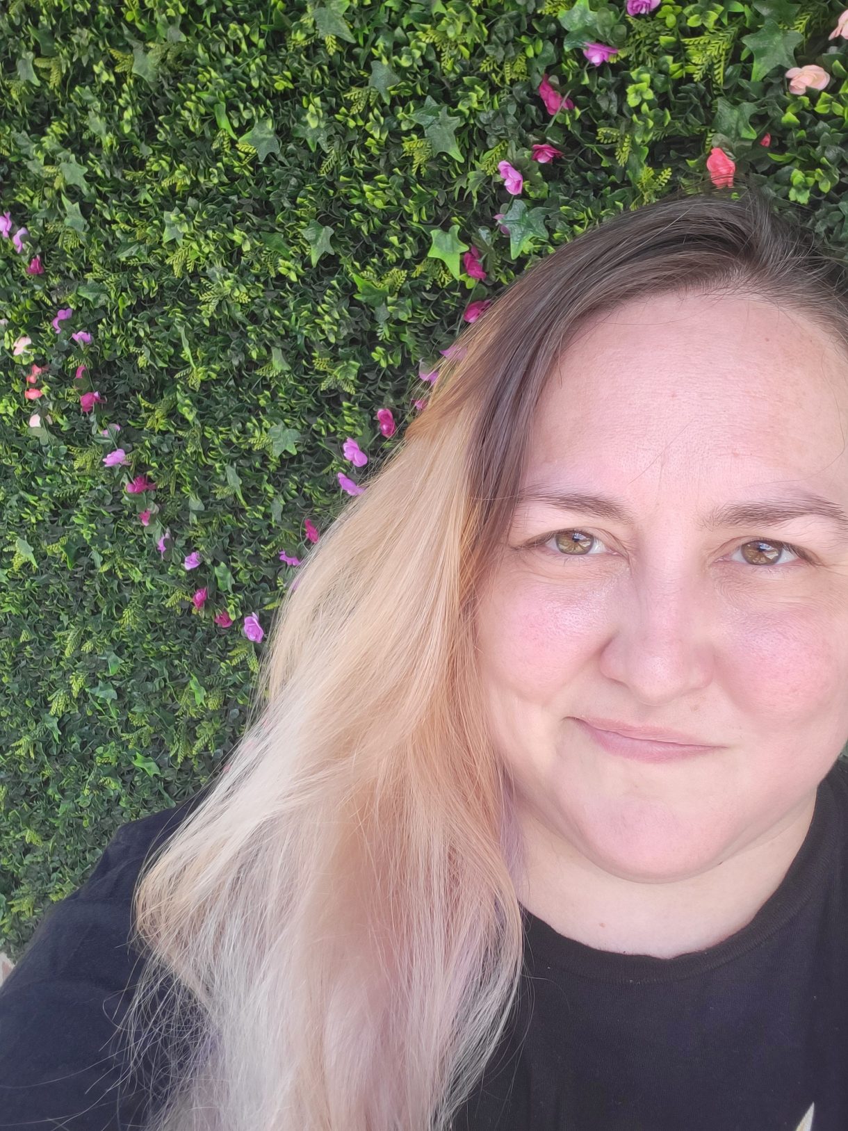 Photo of Valerie Anne a white nonbinary human with long blonde hair smiles up at the camera in front of greenery